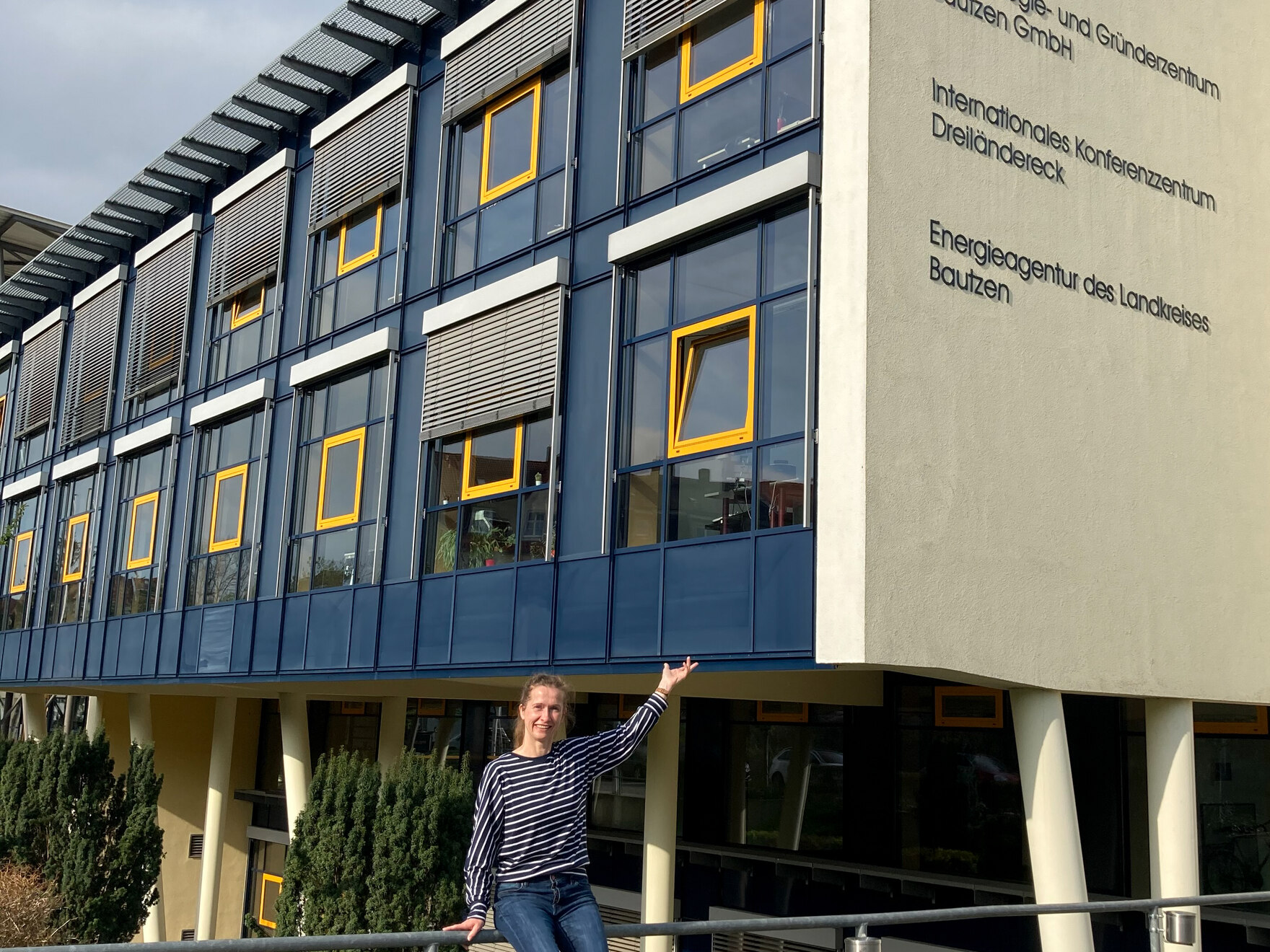 Eine junge Frau sitzt vor einem blauen Haus und zeigt stolz drauf