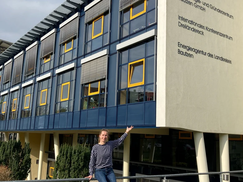 Eine junge Frau sitzt vor einem blauen Haus und zeigt stolz drauf
