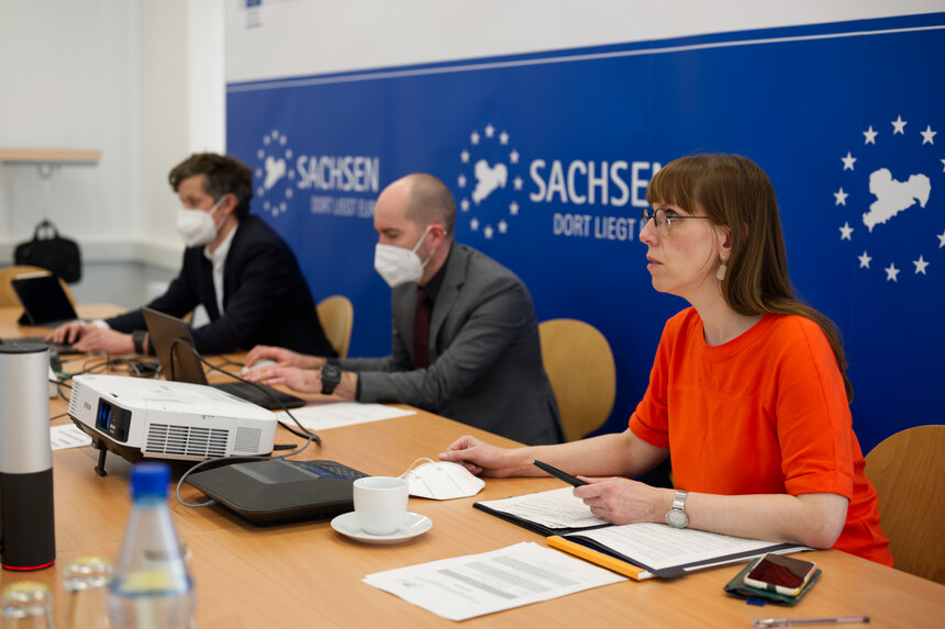 Staatssekretär, Abteilungsleiter Europa und die Ministerin sitzen an einem Tisch und schauen auf ein Bildschirm